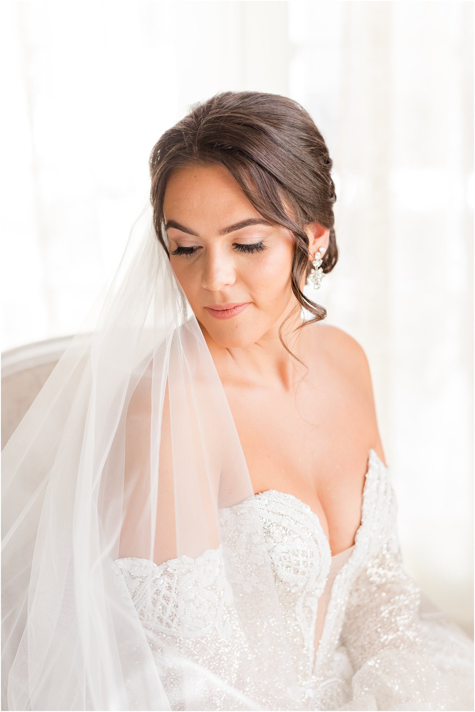 bride sits in tan chair looking over shoulder with veil wrapped around her