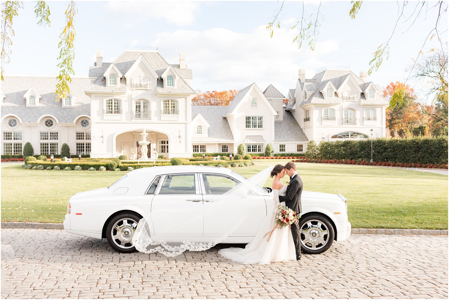 bride and groom pose by white car outside Park Chateau Estate with bride's veil floating
