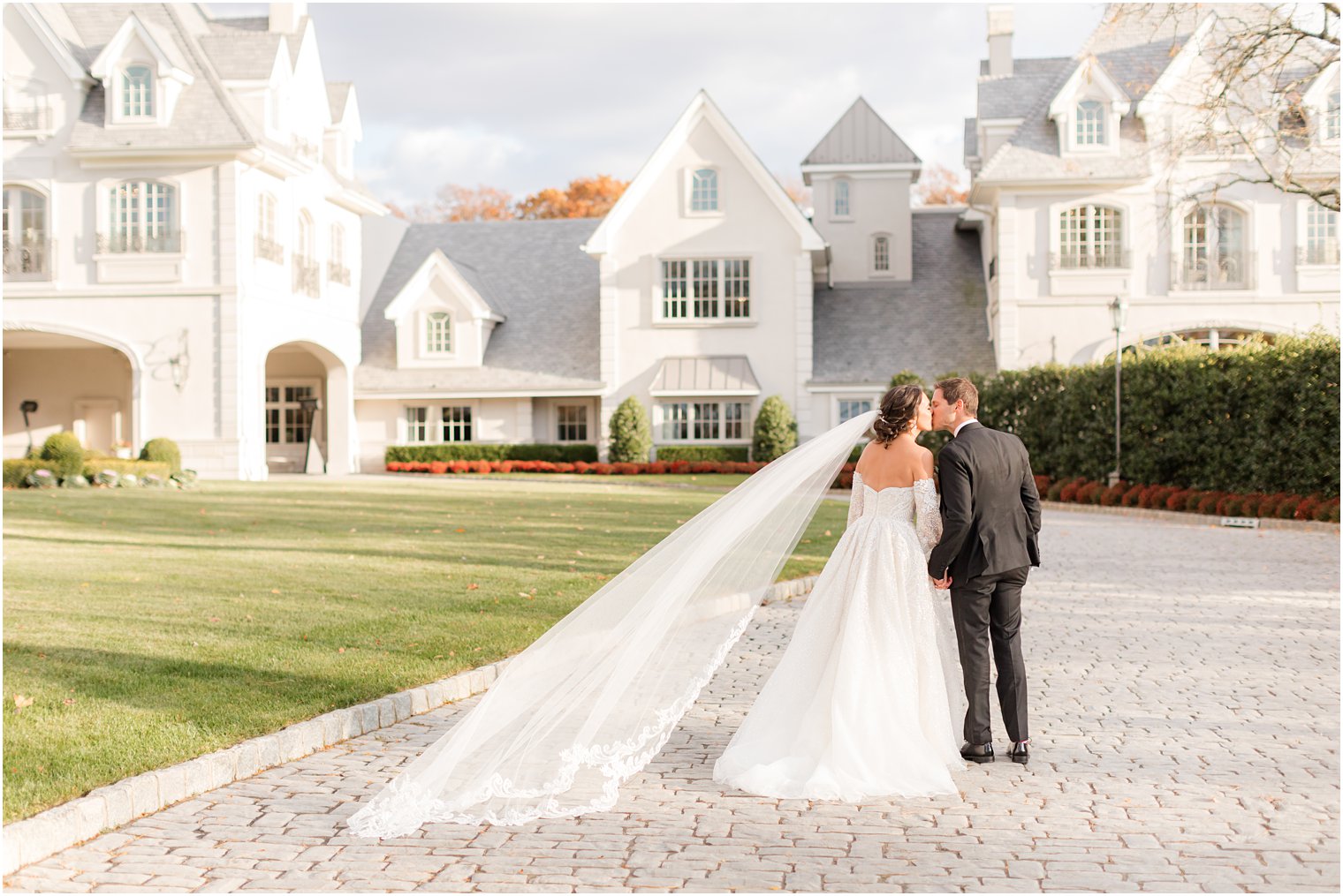 couple holds hands walking down driveway with bride's veil floating behind her 