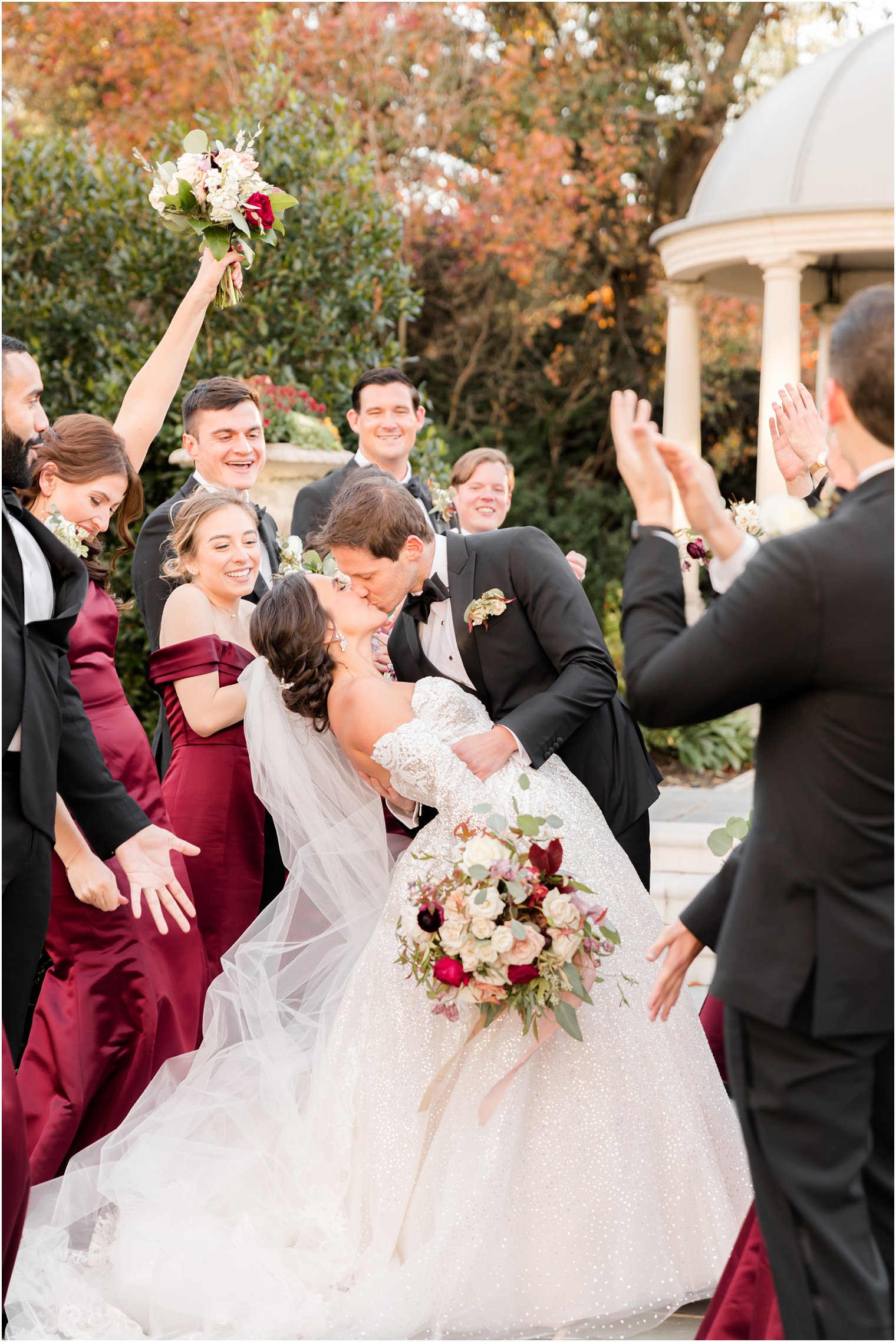 bride and groom kiss surrounded by bridal party in burgundy dresses and black suits