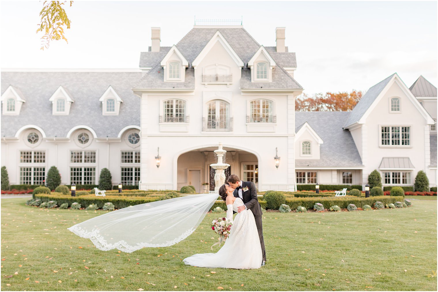 newlyweds kiss on front lawn by fountain at Park Chateau Estate with bride's veil floating behind them