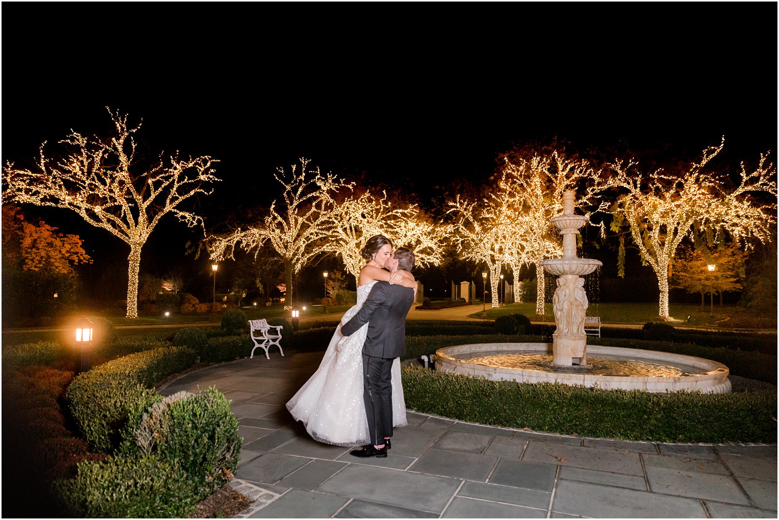 groom lifts bride up outside Park Chateau Estate with fountain and trees with holiday lights