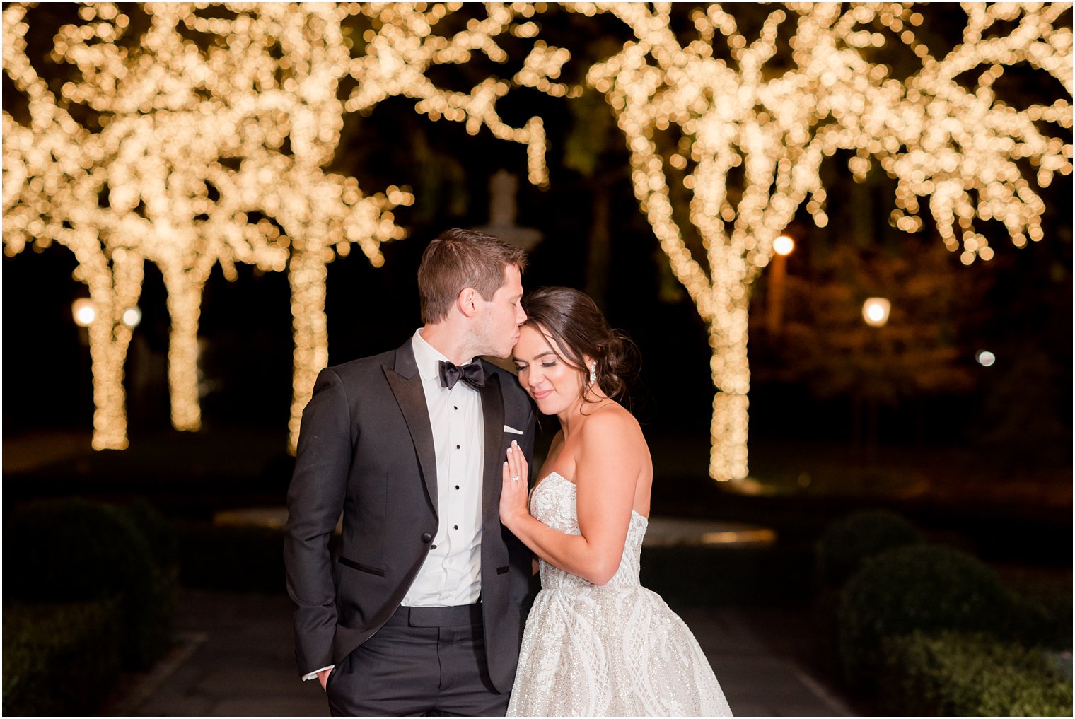 groom kisses bride outside Park Chateau Estate at night with trees in Christmas lights