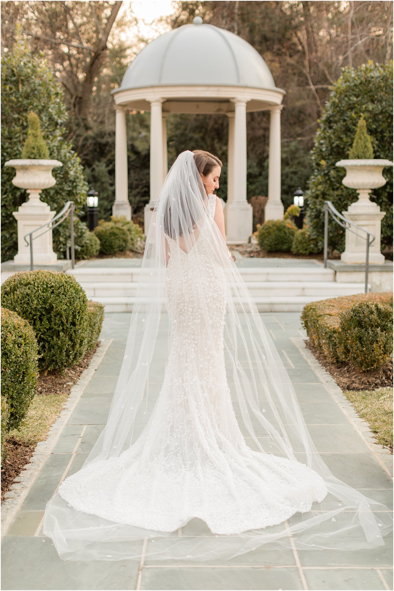 bride stands in gardens showing off back of wedding gown with veil draped behind her 