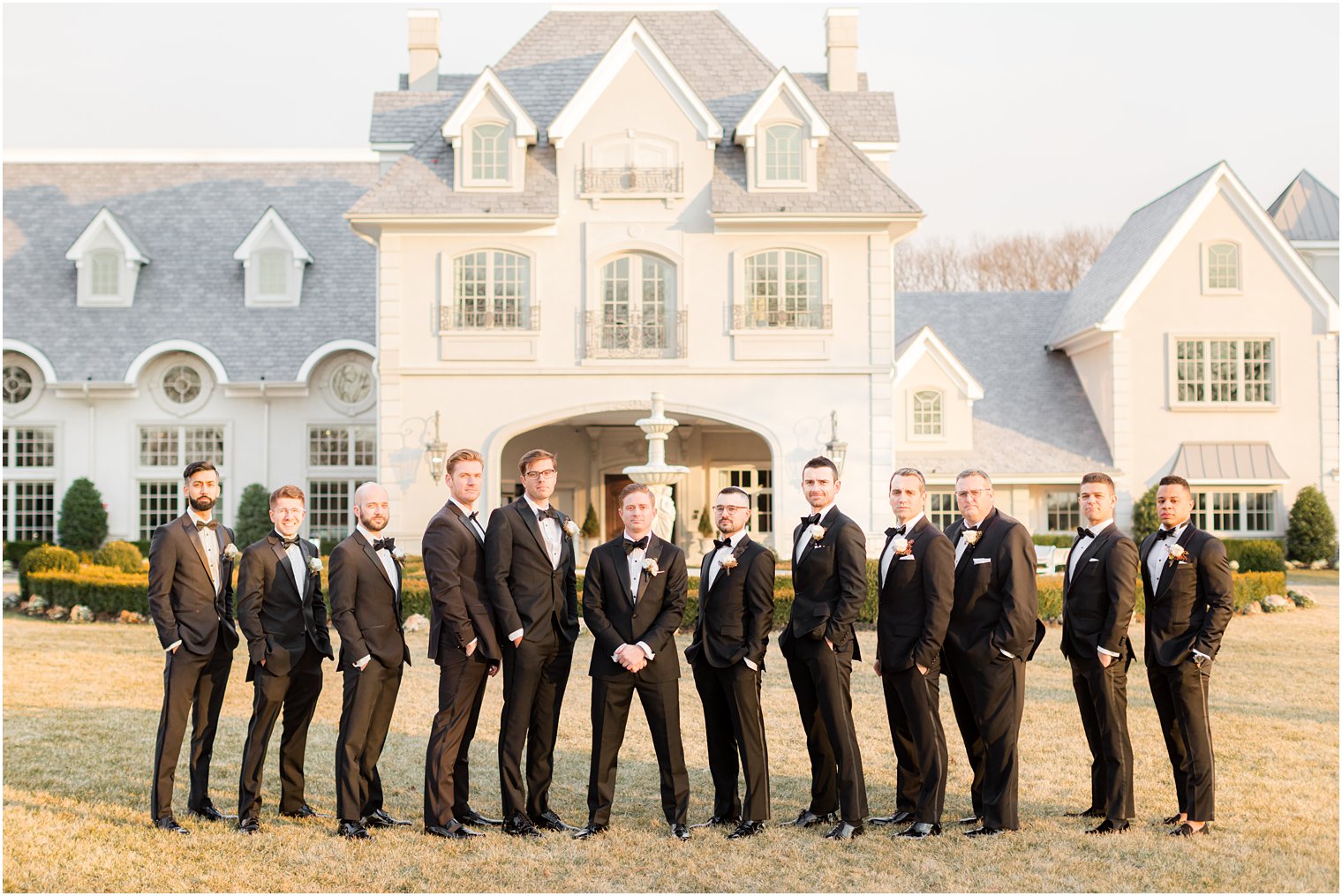 groom stands on lawn at Park Chateau with groomsmen with hands in pockets