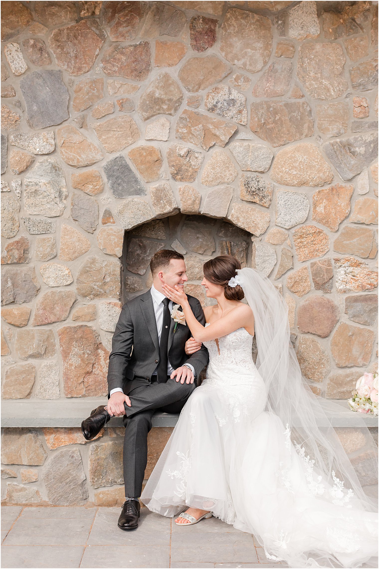 newlyweds sit by stone fireplace at The Refinery at Perona Farms