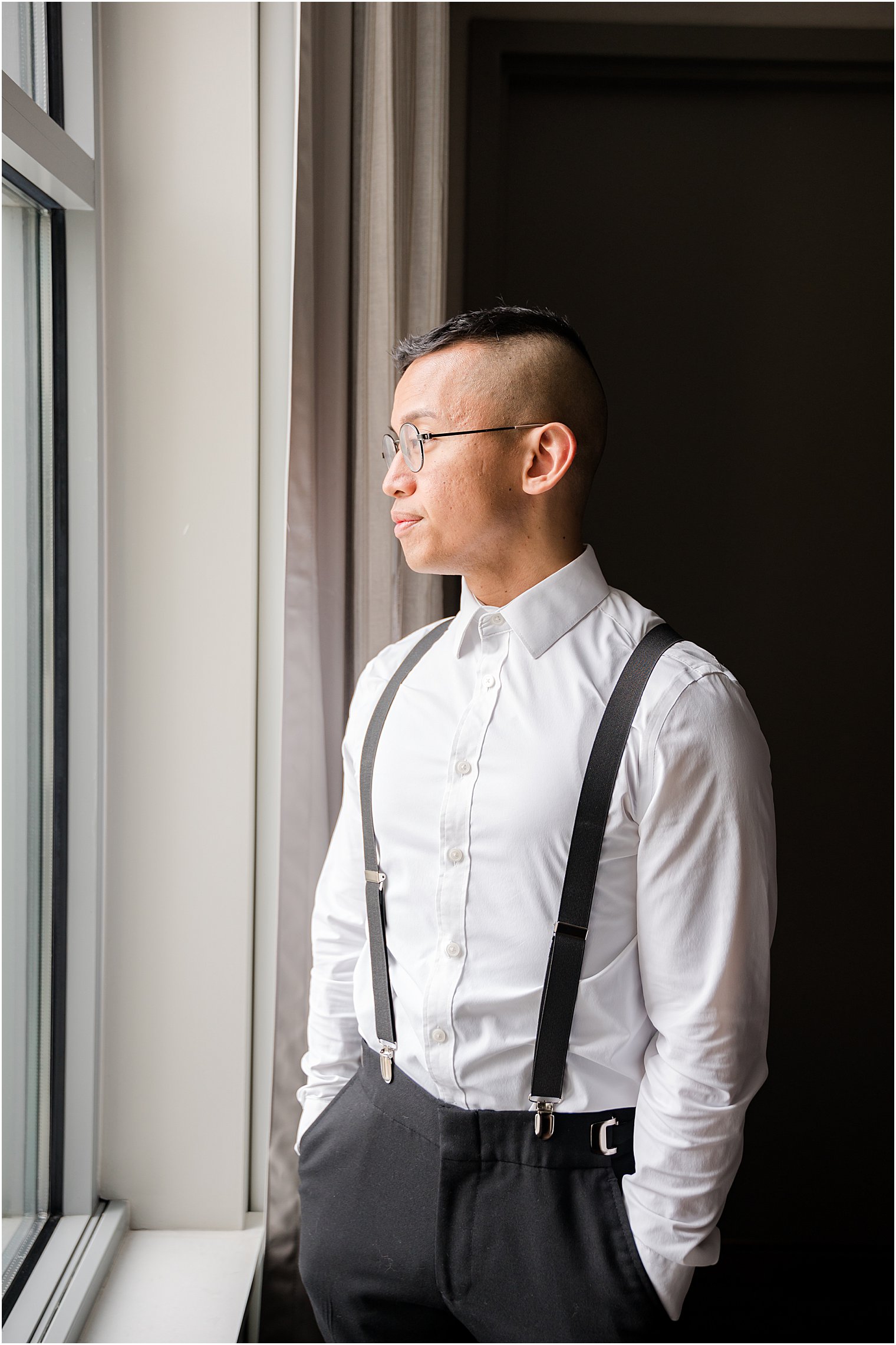 groom looks out window in long sleeves and suspenders