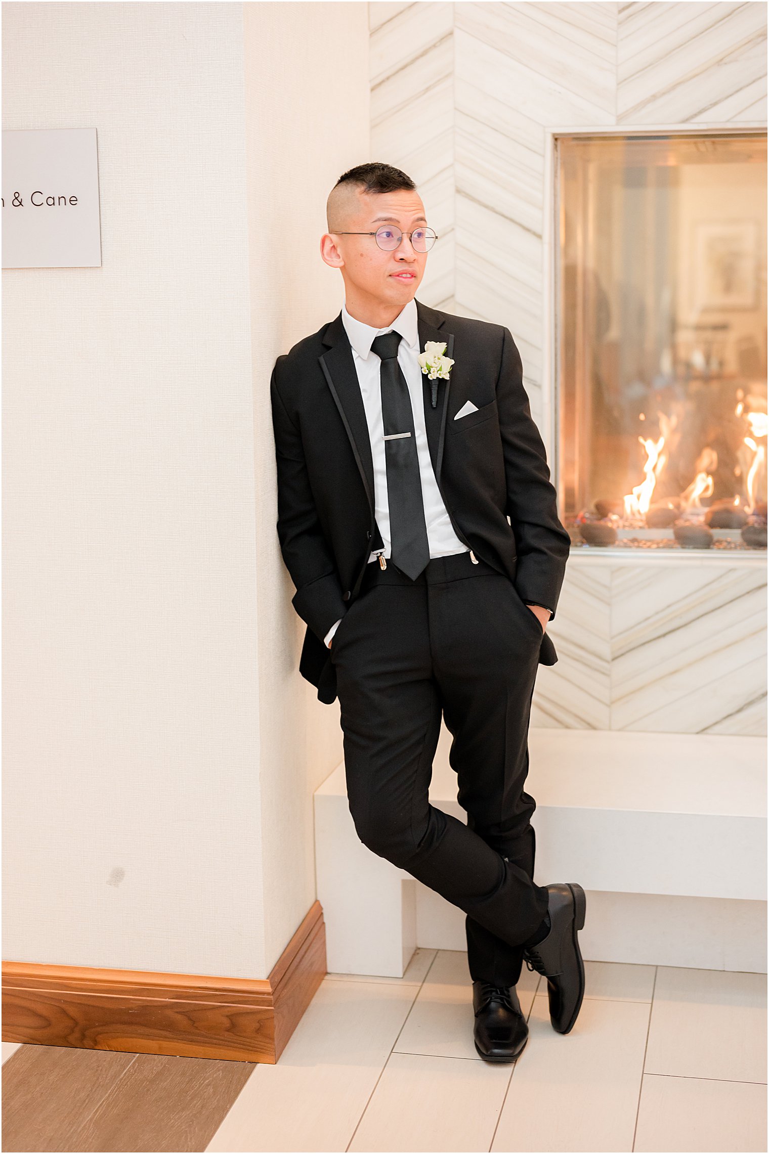 groom leans against white wall by fireplace