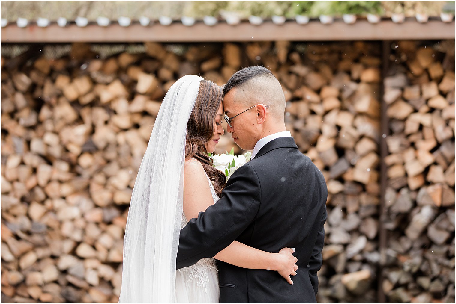 bride and groom stand with foreheads touching by woodpile 