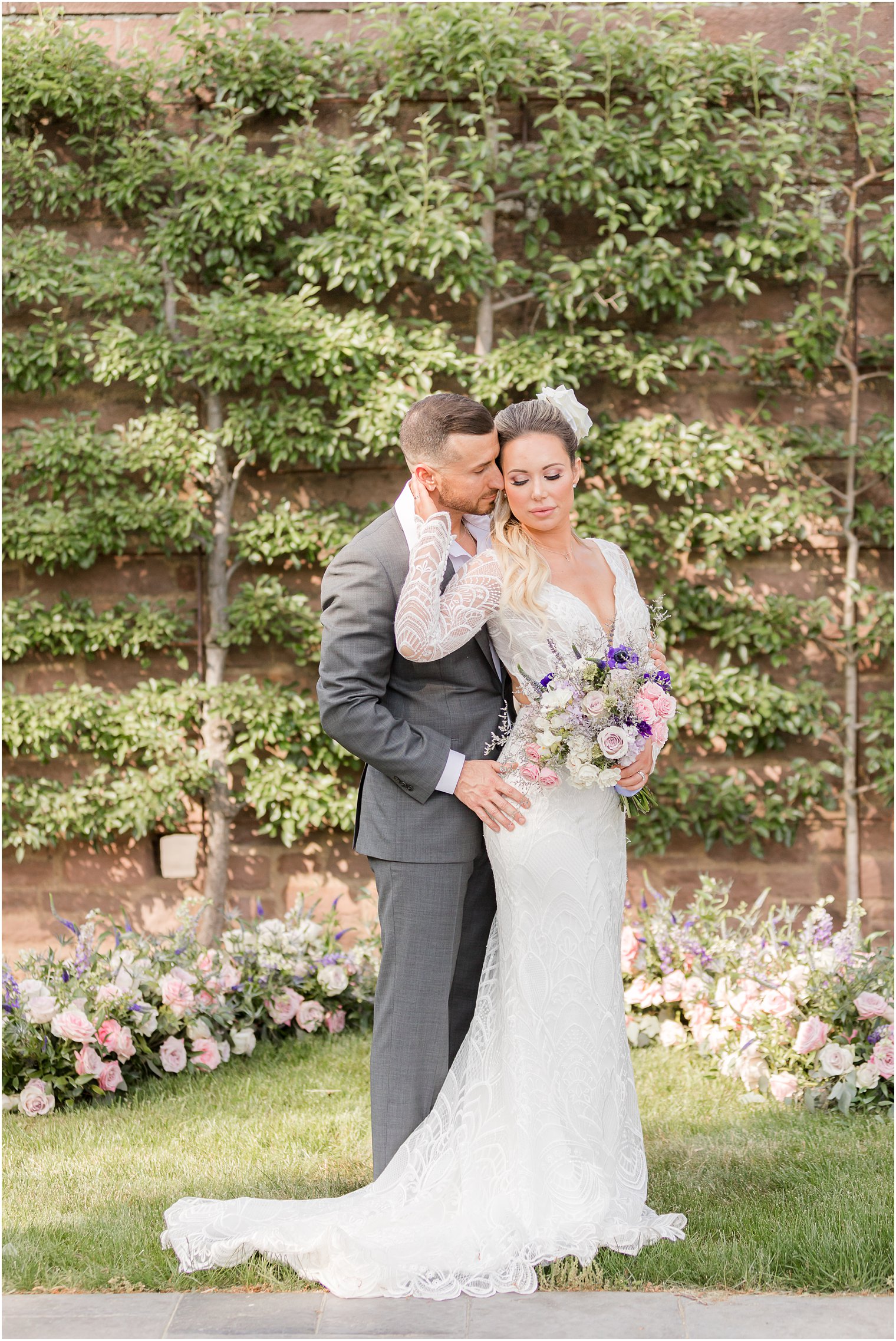 groom stands with bride in front of him by brick wall at Tyler Gardens