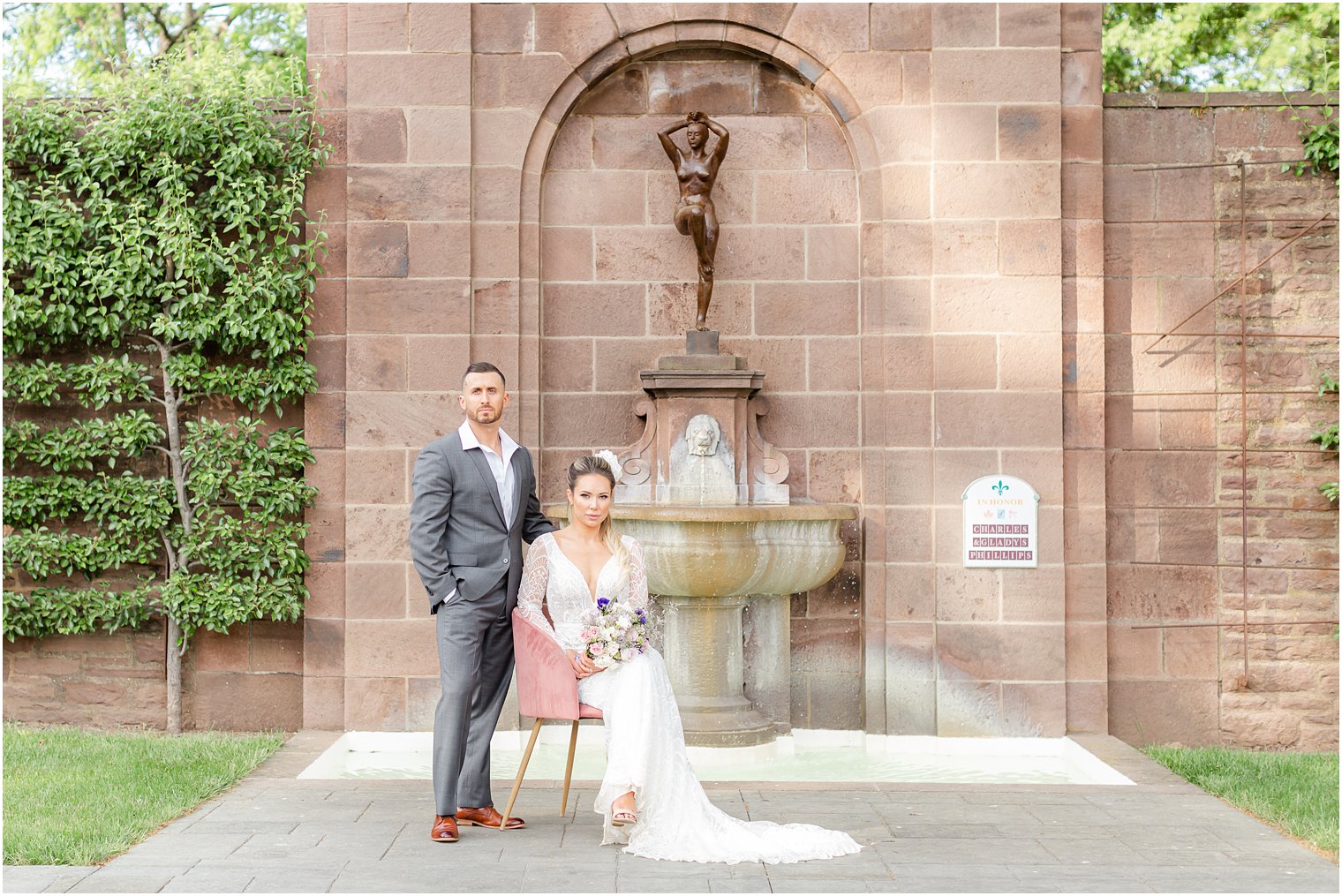groom stands by bride sitting in pink chair by brick fountain inside Tyler Gardens