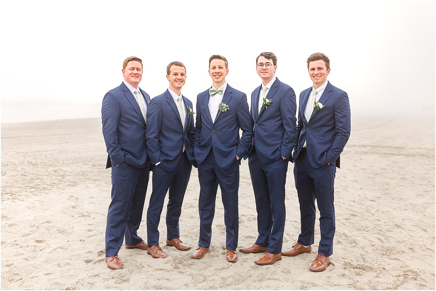 groom stands with groomsmen in navy suits on foggy beach