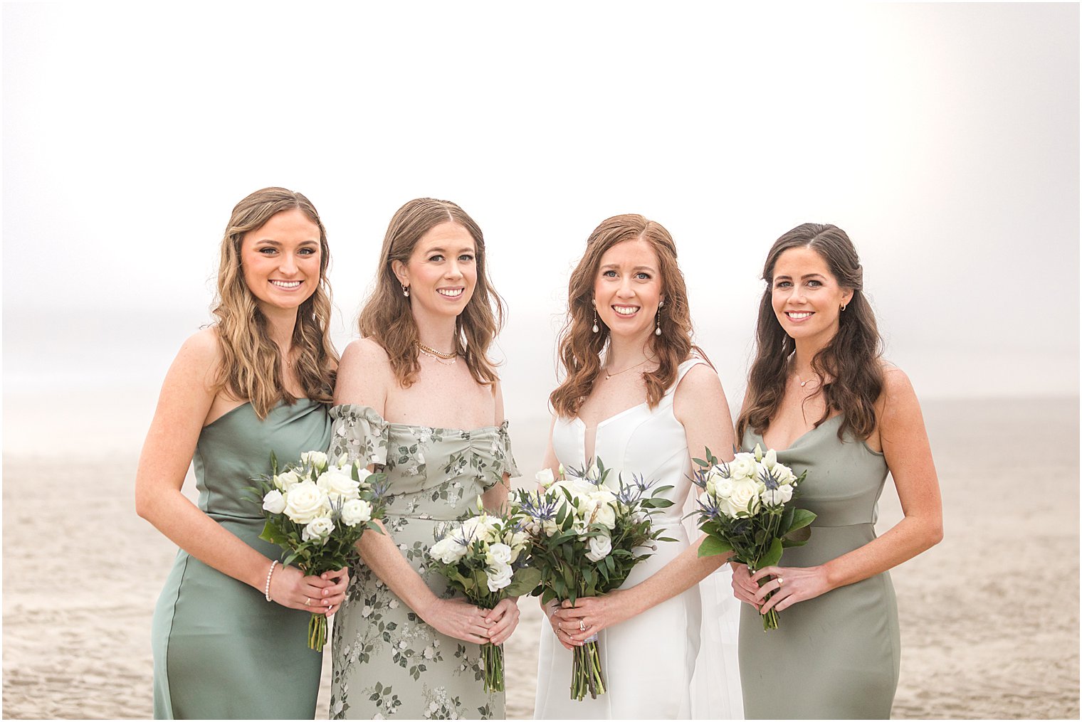 bride and bridesmaids in mismatched green gowns stand on beach