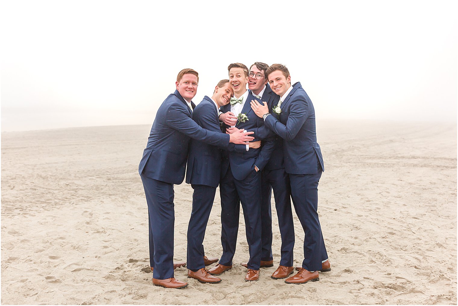 groomsmen in navy suits hug groom on foggy beach