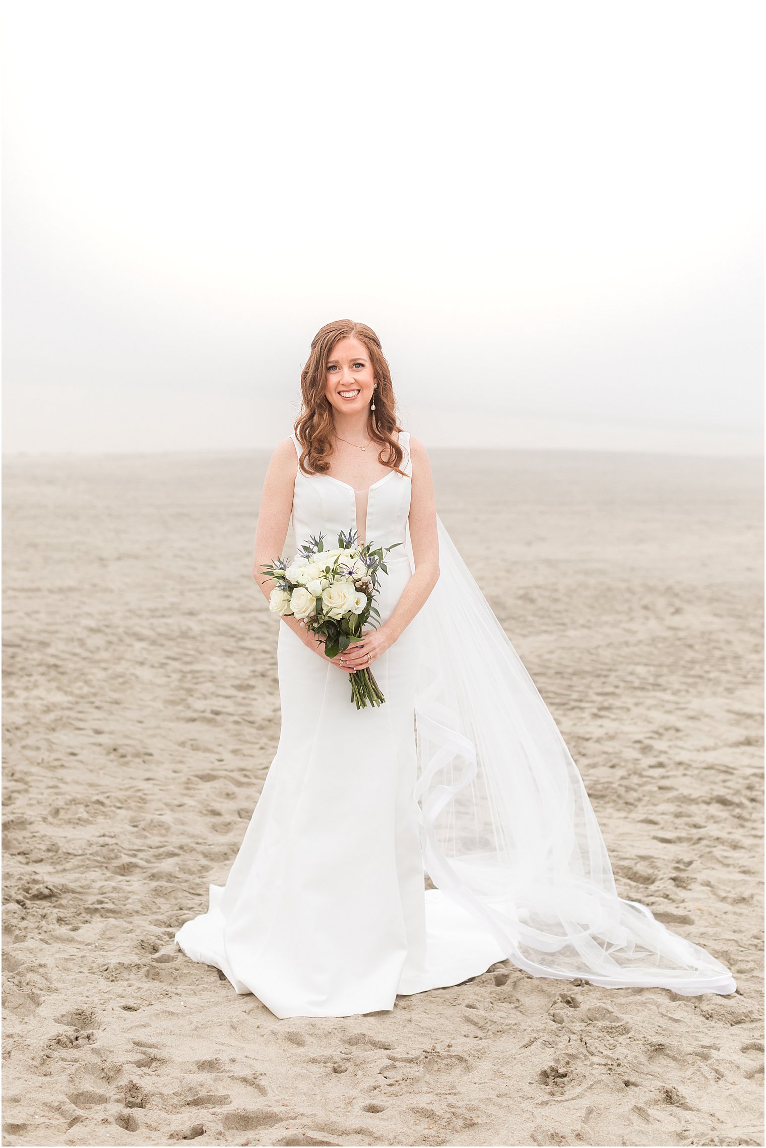 bride stands on foggy beach with bouquet of white flowers