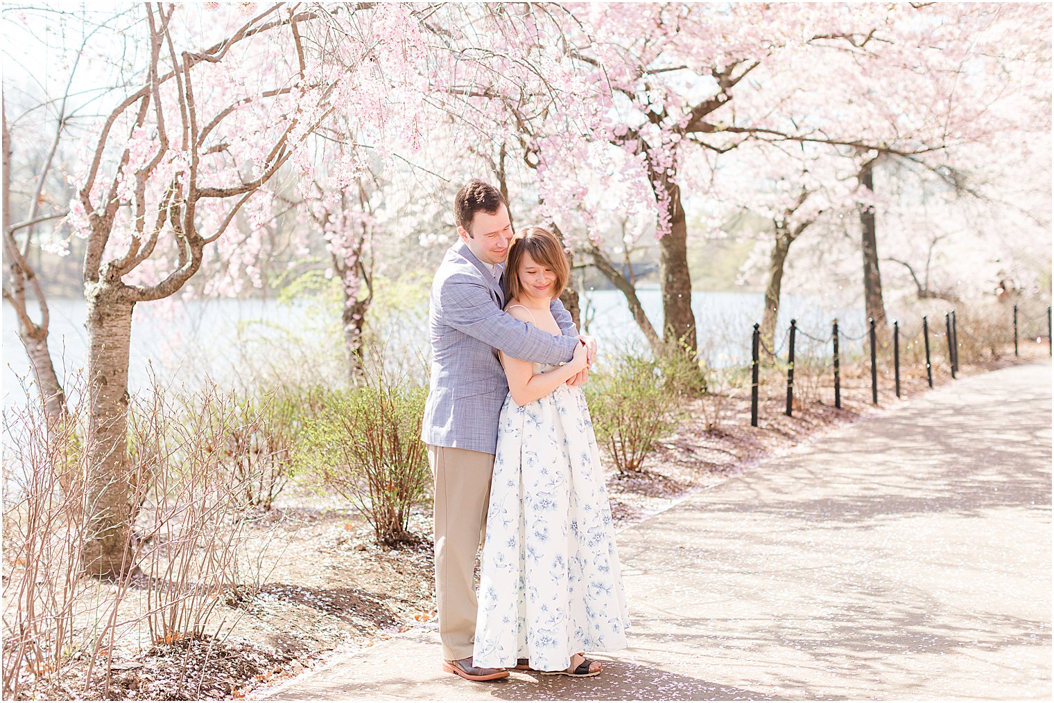 man hugs fiancee under cherry blossom trees at Branch Brook Park