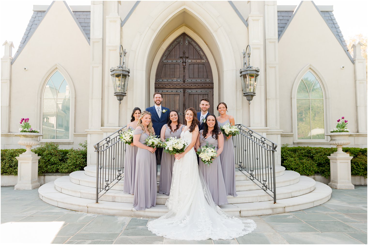 bride stands on steps of chapel with bridal party in pastel gowns