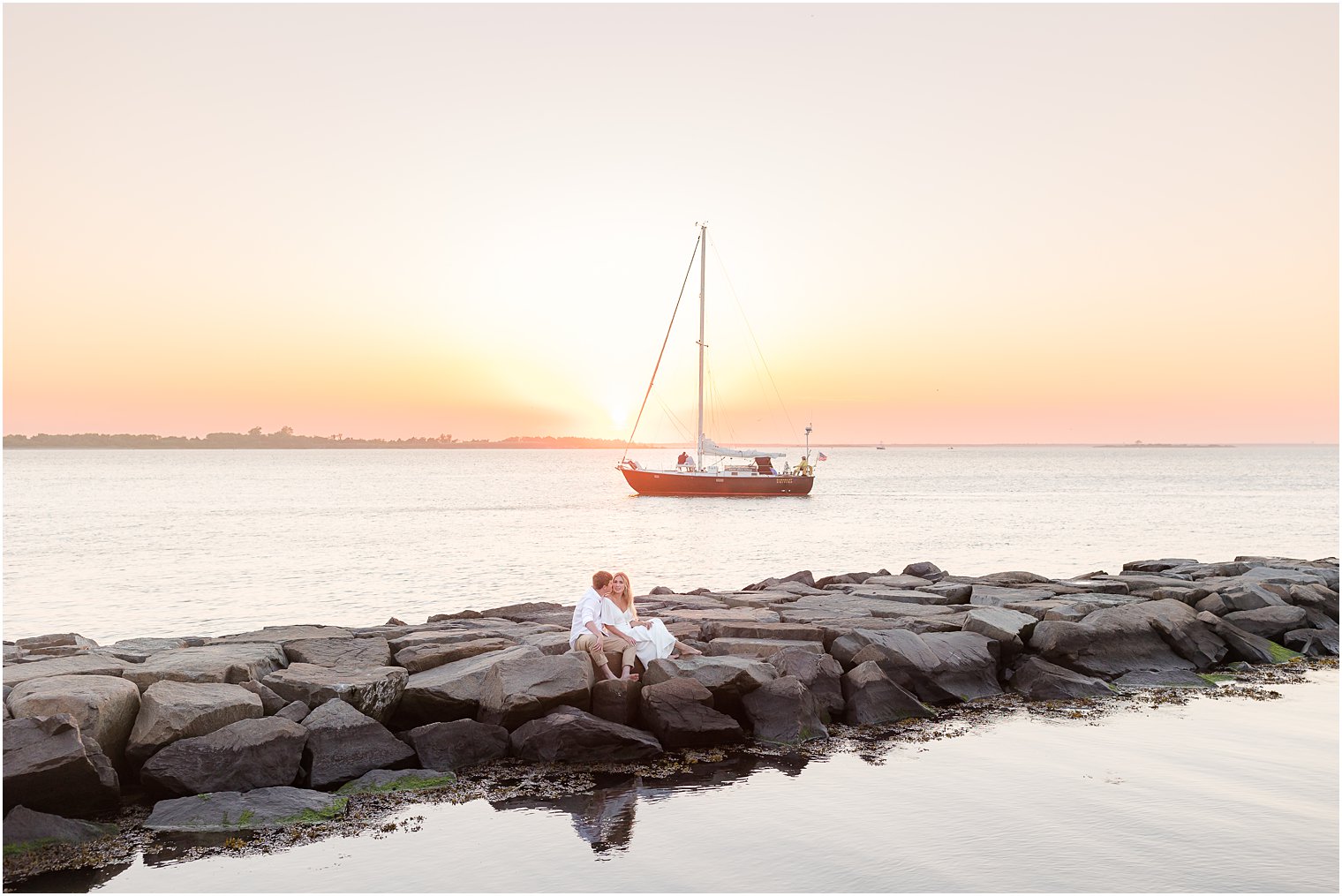 engaged couple sit on rocks with sailboat behind them at Long Beach Island