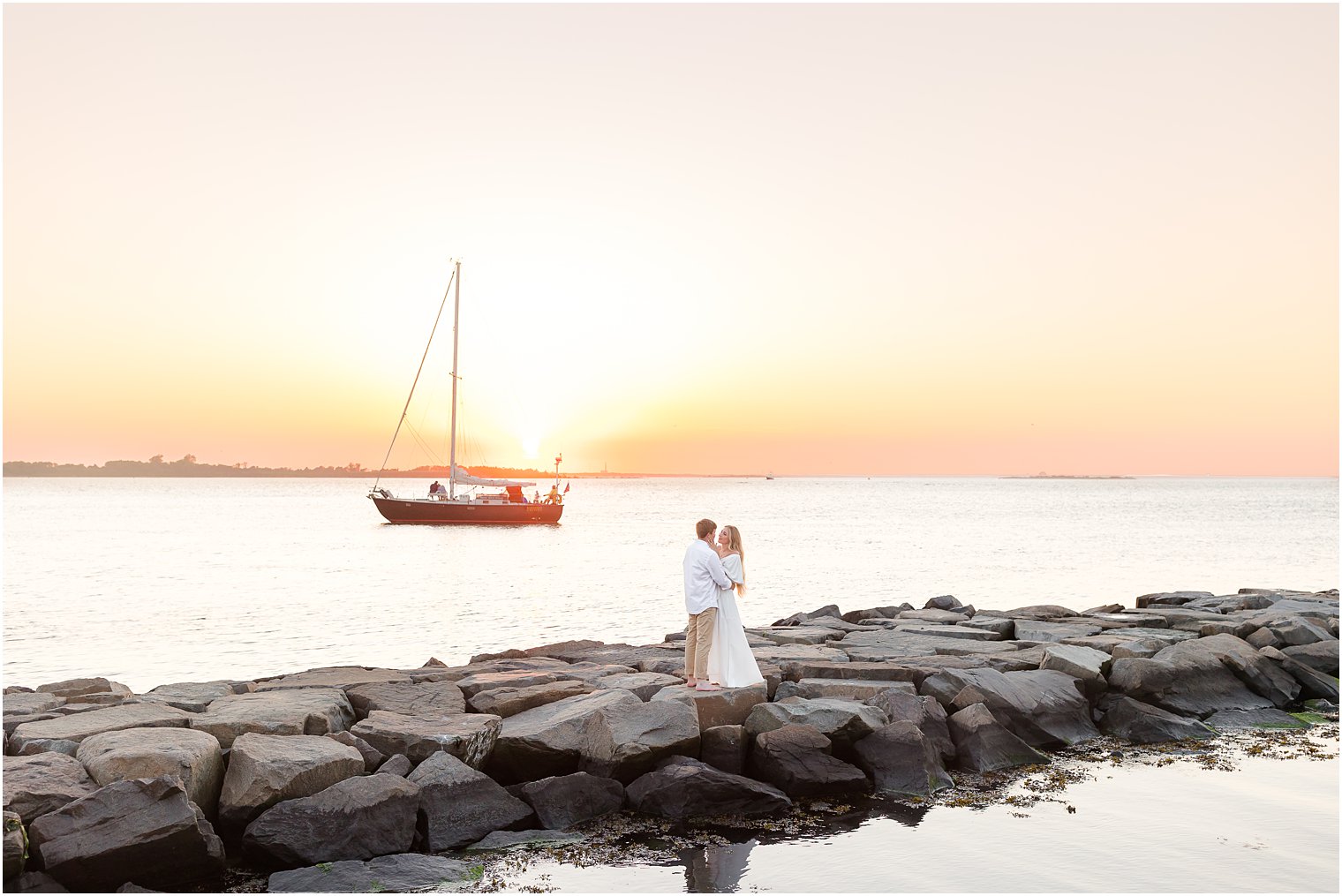 couple hugs while sailboat drives by during Long Beach Island engagement session