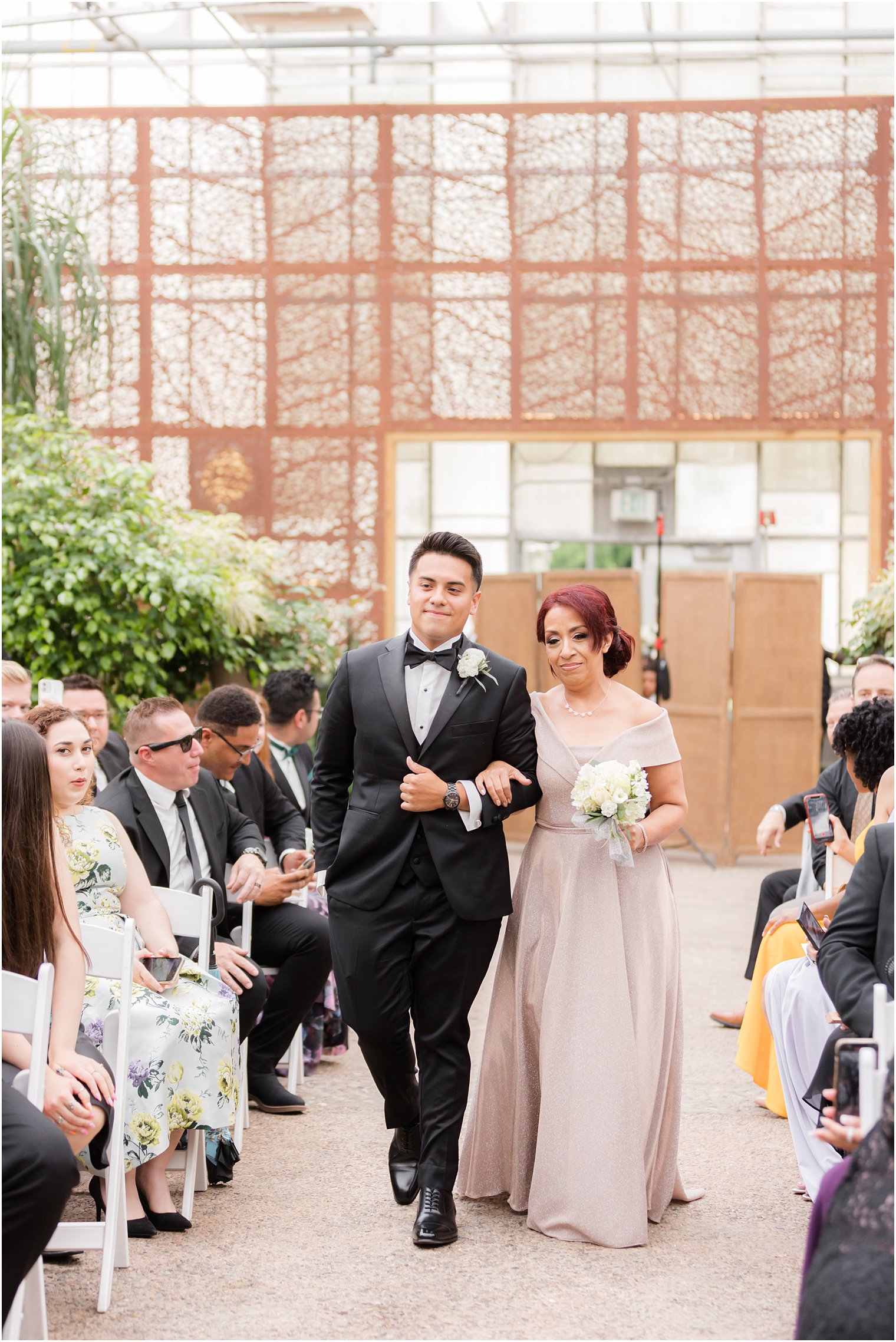 groom walks mother in pink gown down aisle for Fairmont Park Horticulture Center wedding ceremony in greenhouse