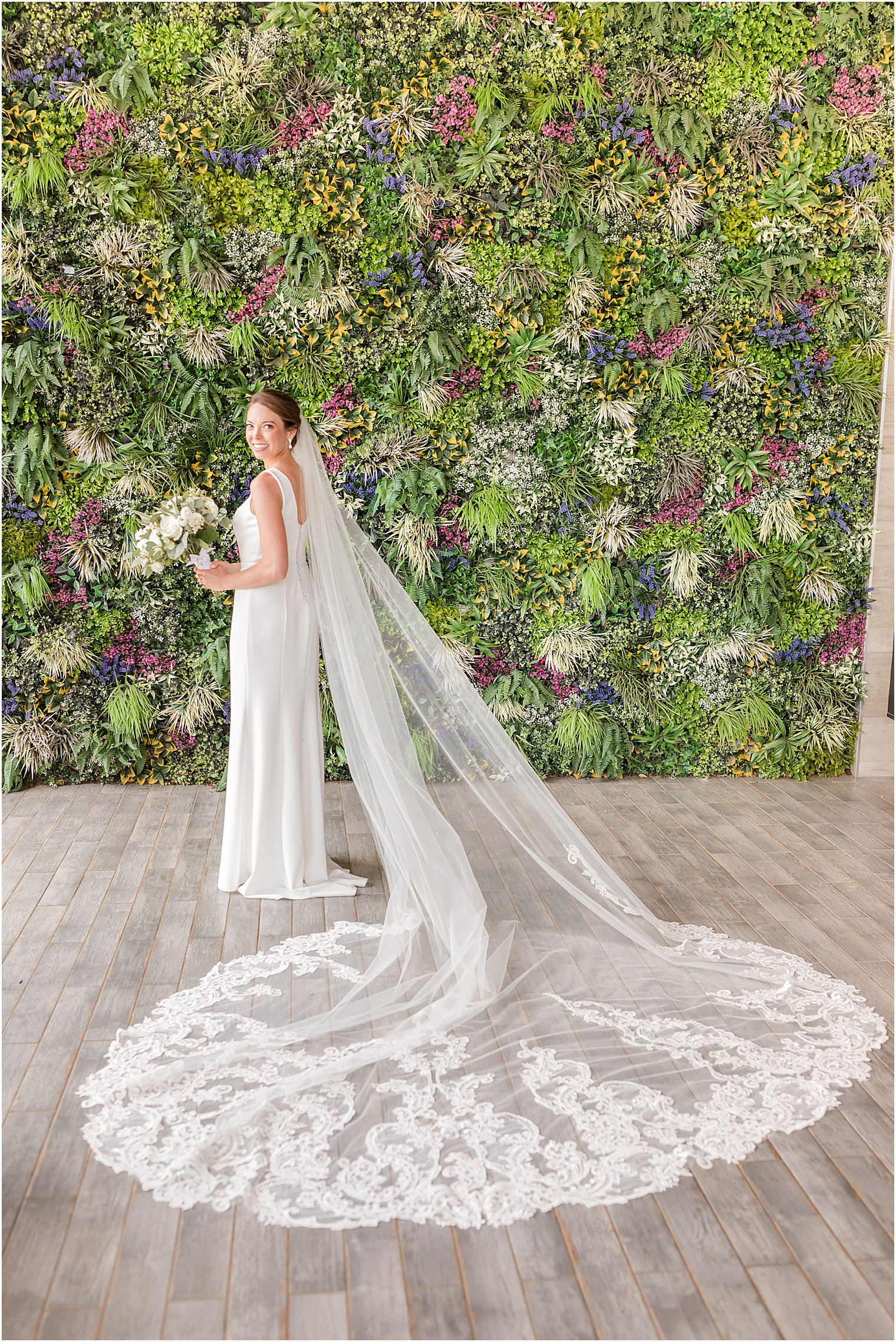 bride stands by greenery wall with wedding dress and veil behind her