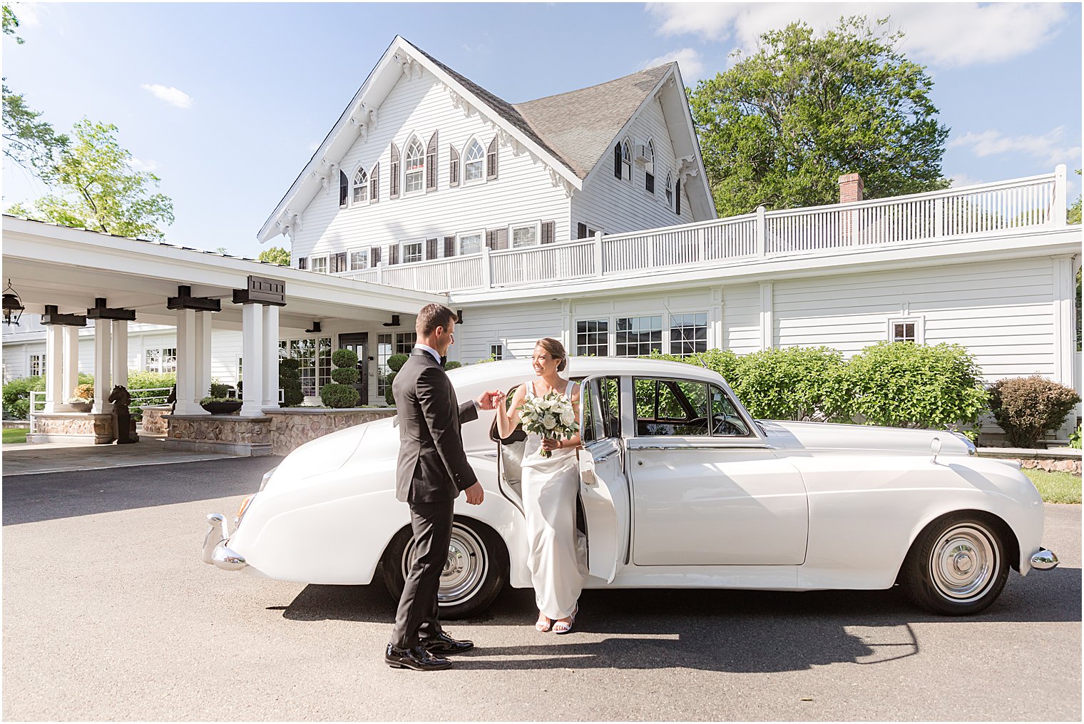 groom helps bride get out of white classic car in front of Ryland Inn