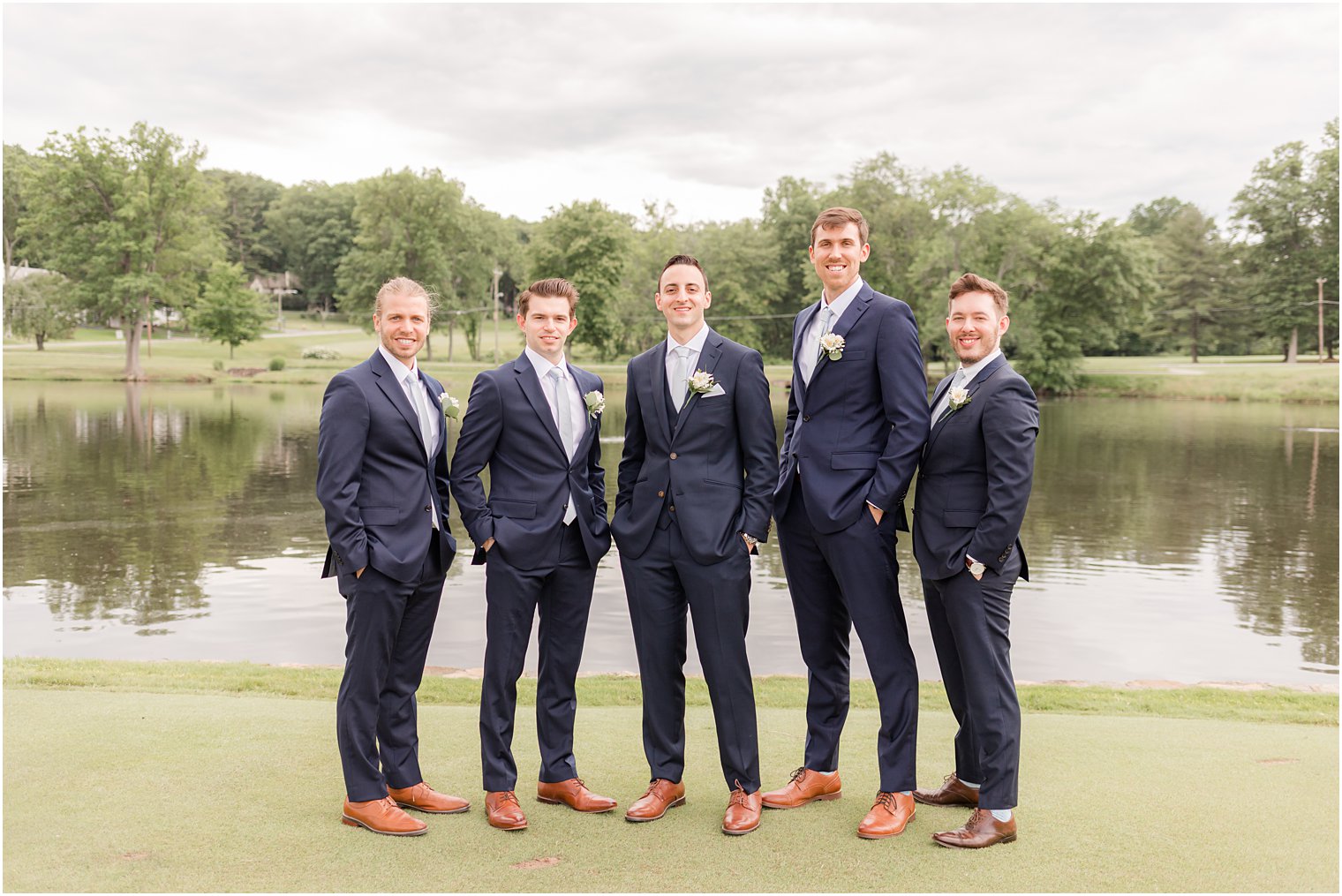 groom stands with groomsmen in navy suits by lake at Spring Brook Country Club