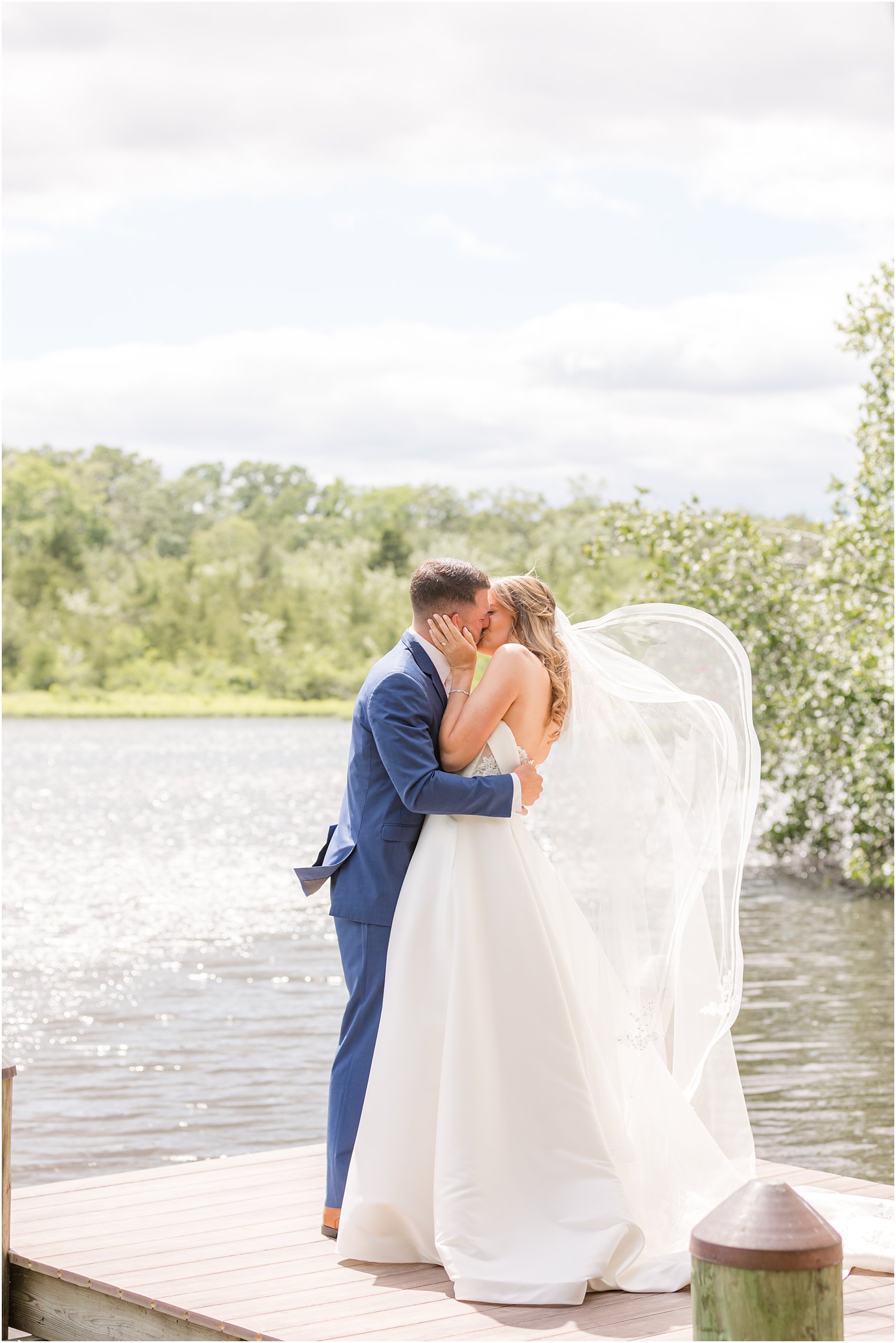 bride and groom kiss on dock with veil floating behind her