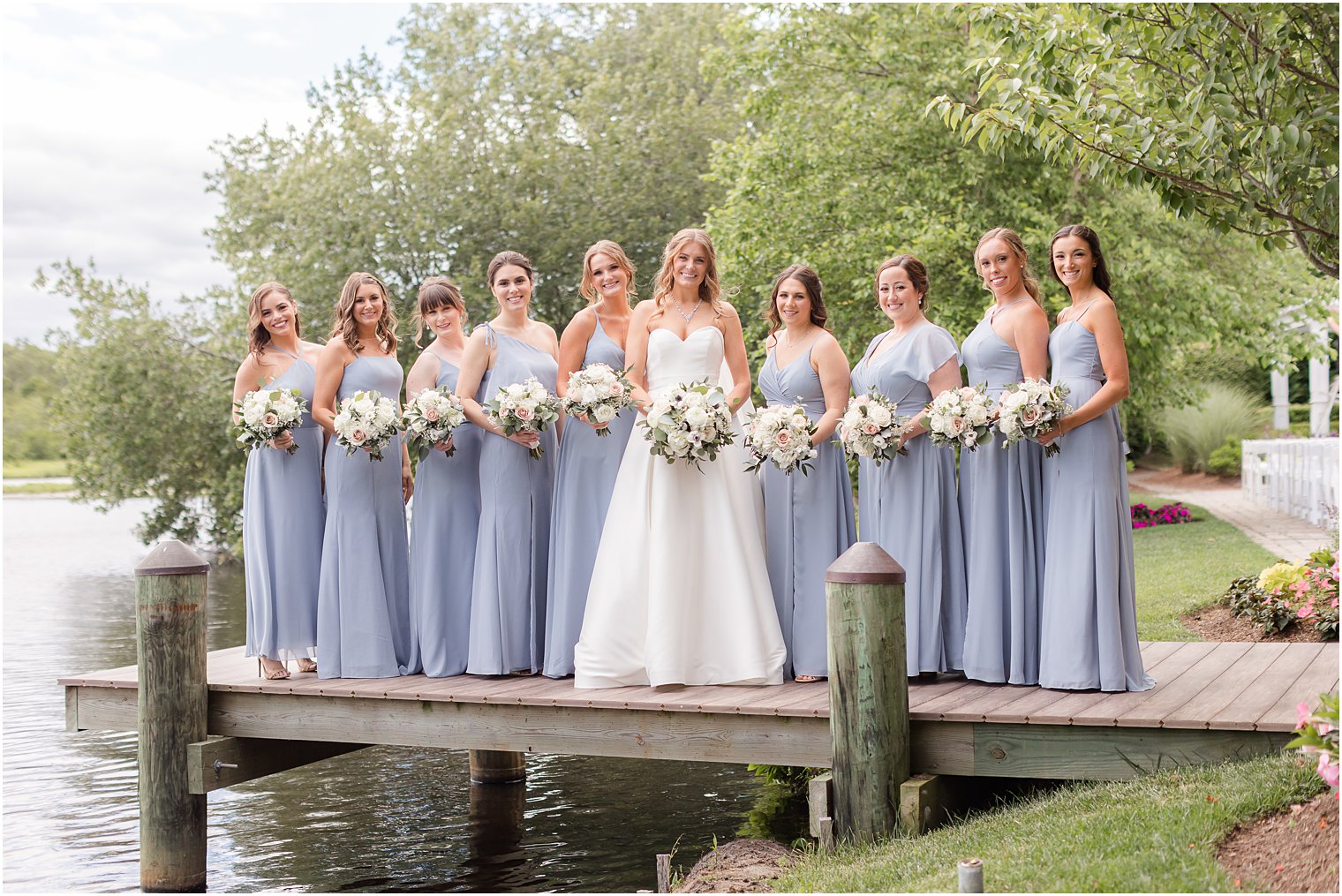 bride stands on dock with bridesmaids in blue dresses