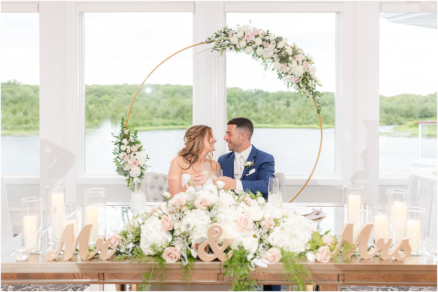 newlyweds sit a sweetheart table by window of The Mill Lakeside Manor