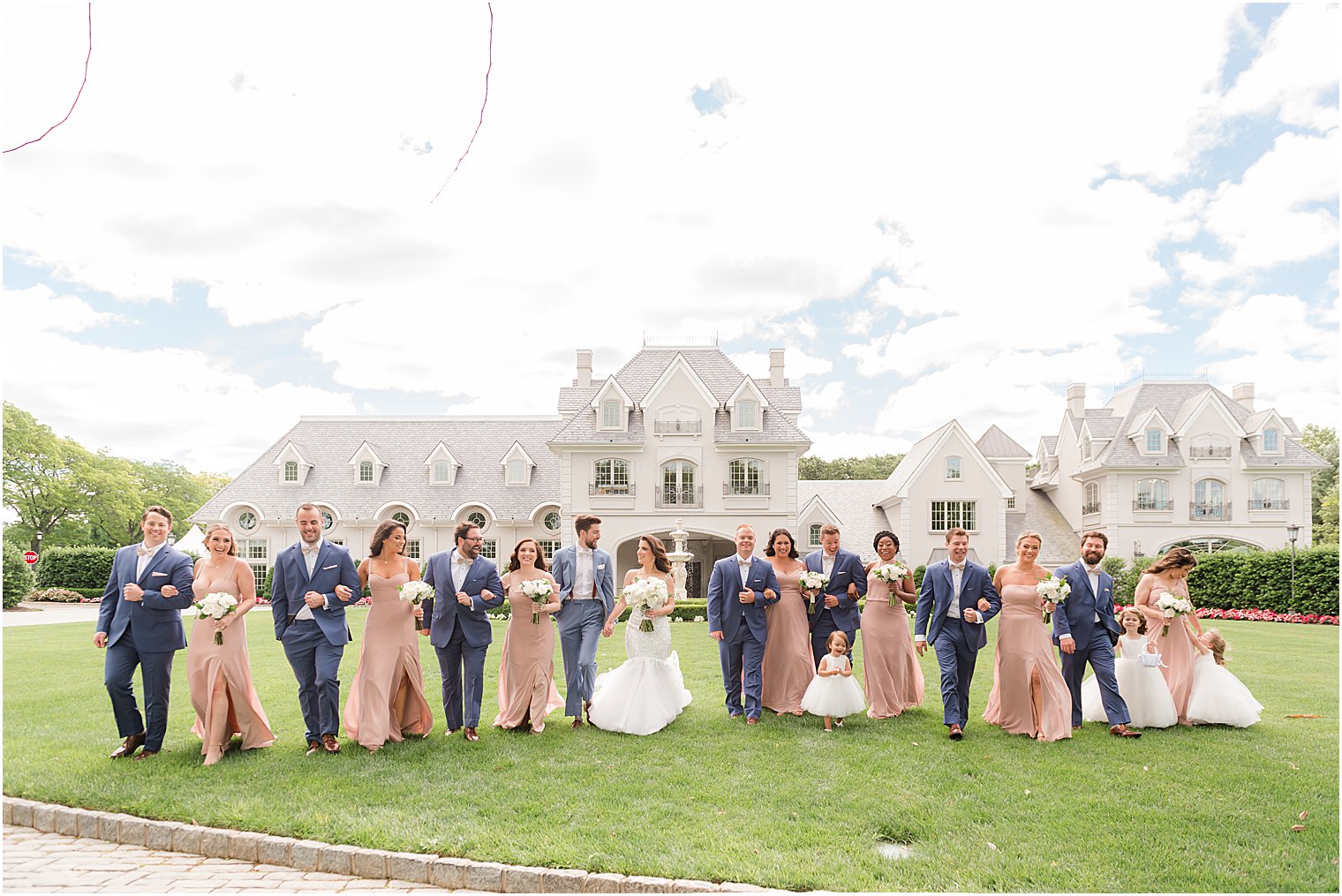 bride and groom stand on lawn of Park Chateau Estate with wedding party in blue and peach