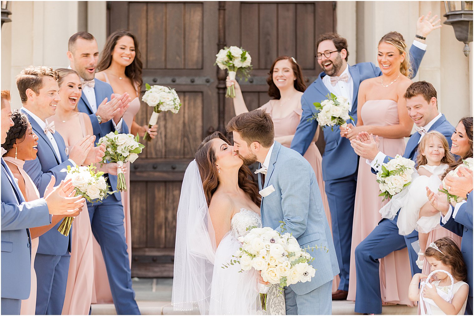 newlyweds kiss while wedding party cheers on steps of Park Chateau Estate chapel
