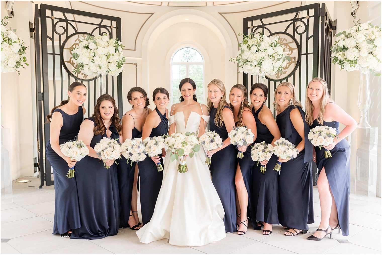 bride stands with bridesmaids in navy gowns with ivory and pale pink bouquets