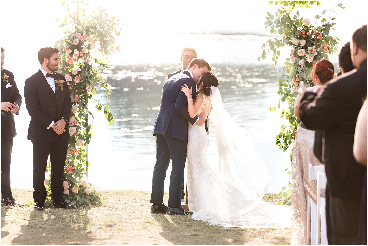 bride and groom kiss between floral arches in outdoor wedding ceremony by water in Franklin Lakes NJ