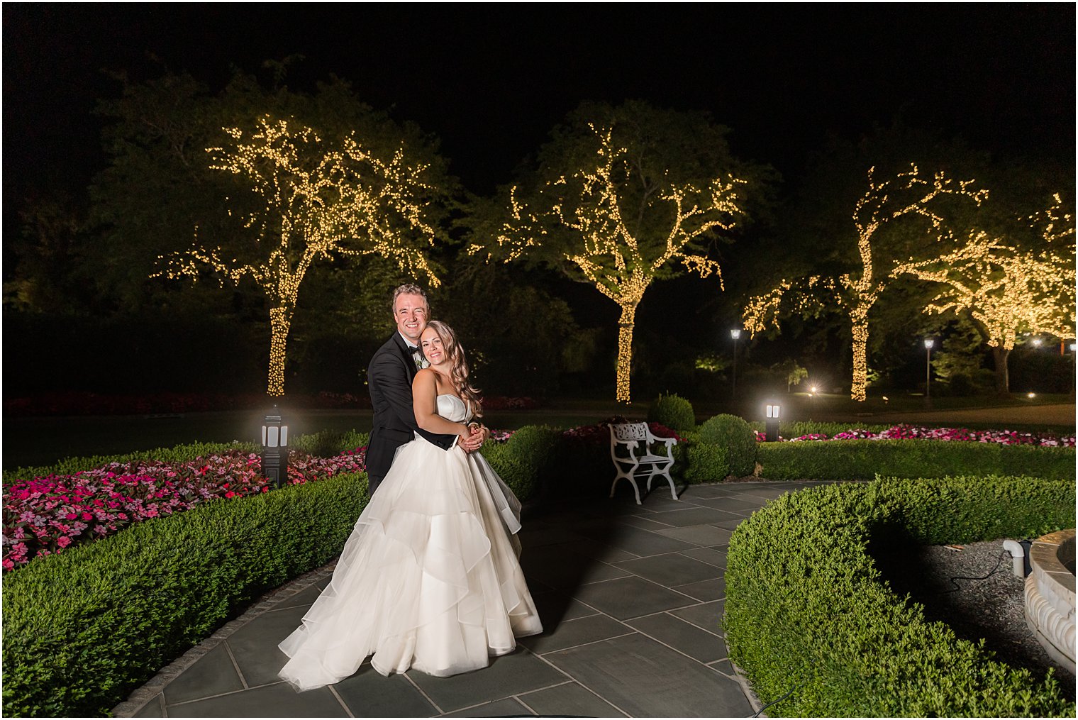 grom hugs bride from behind on sidewalk at night outside Park Chateau Estate