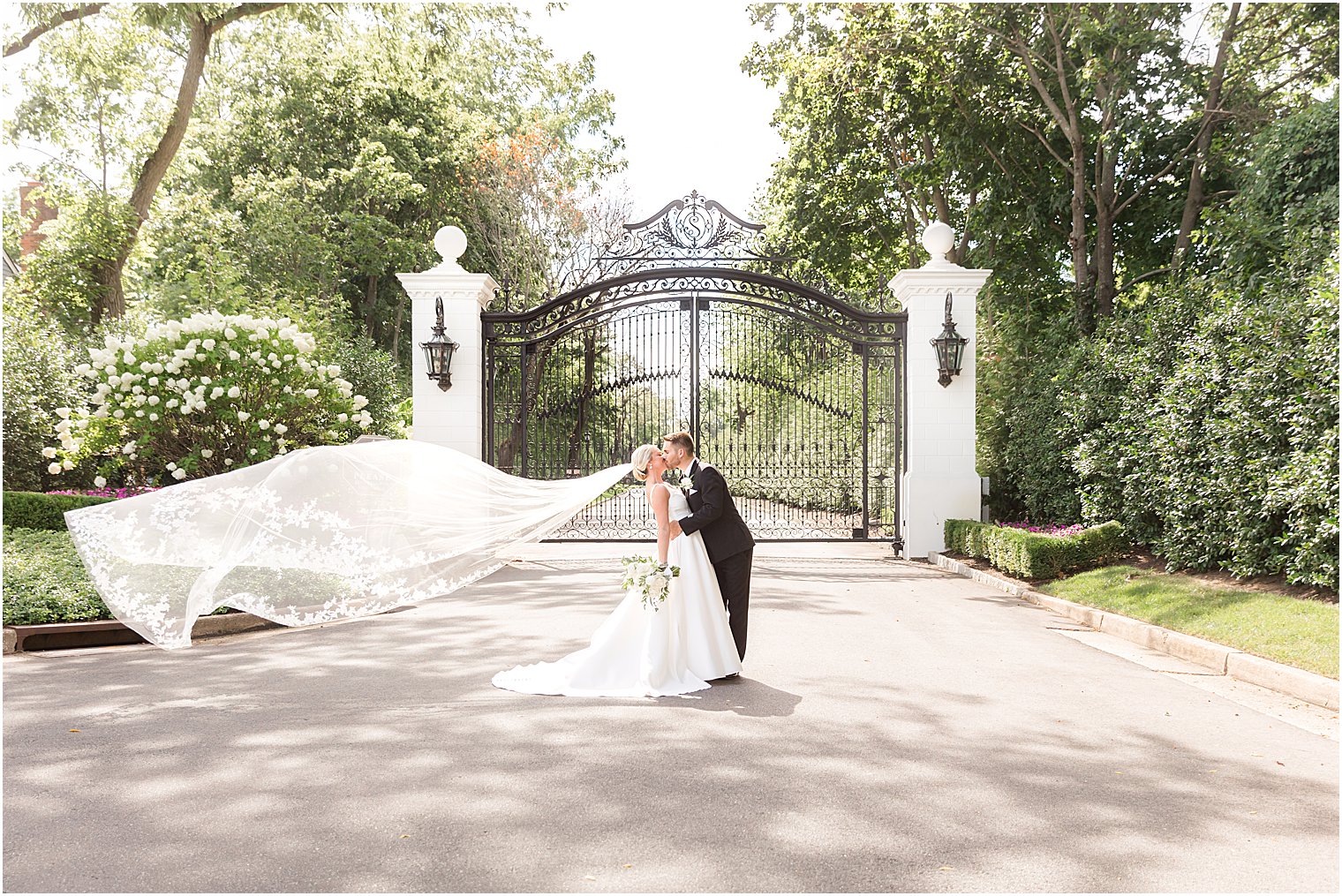 newlyweds kiss by wrought iron gate outside Shadowbrook at Shrewsbury with bride's veil floating behind them