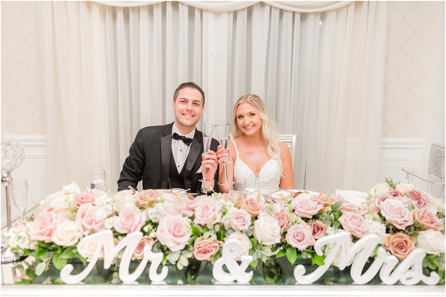 newlyweds toast champagne glasses at sweetheart table behind pink and white flowers