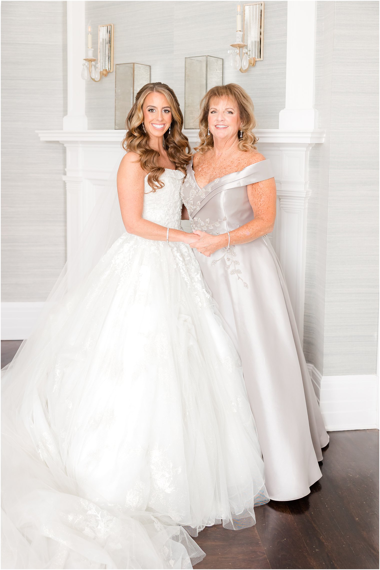 bride stands with mother in silver gown by white fireplace at Shadowbrook at Shrewsbury