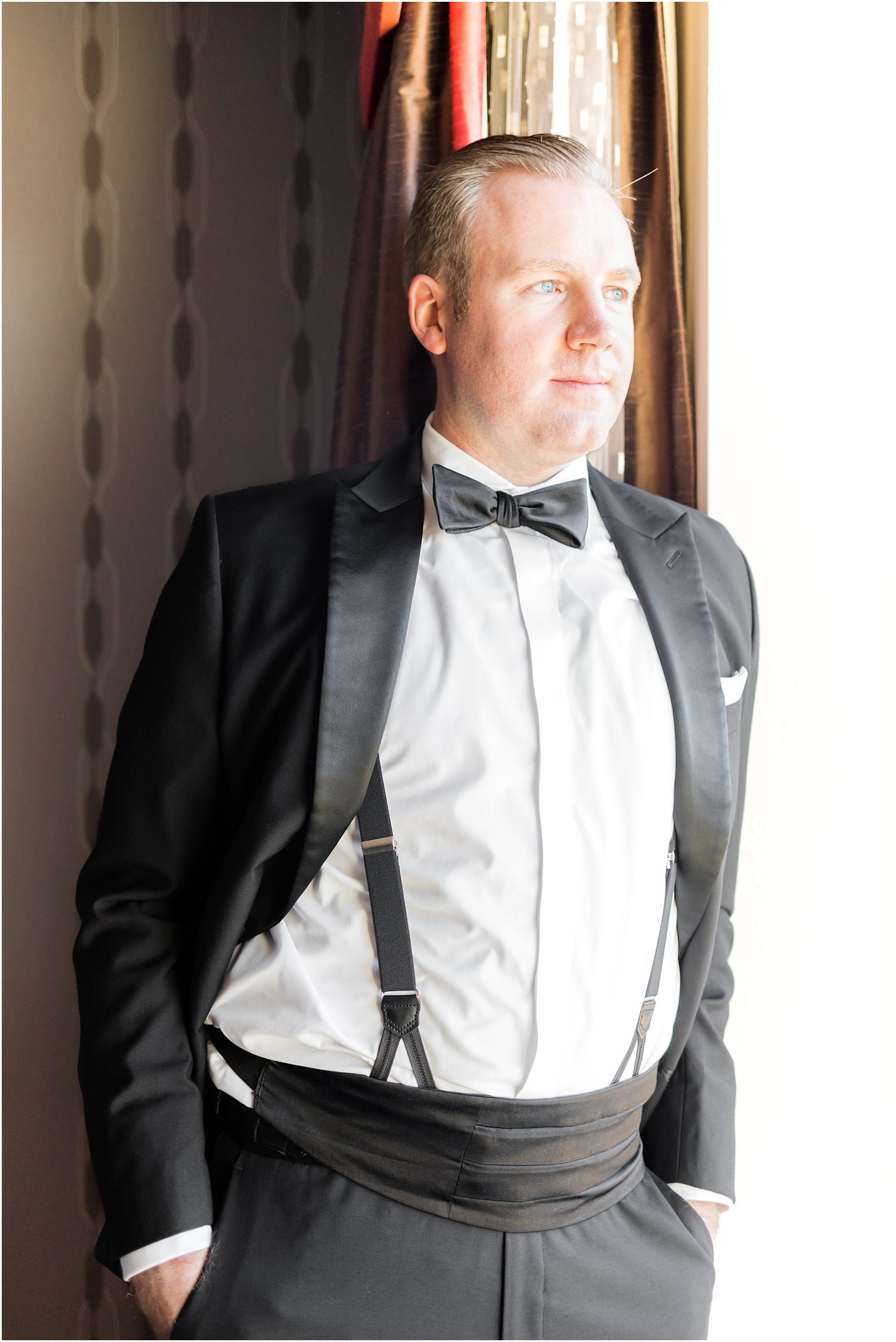 groom stands with hands in tux pockets looking out window in New Jersey 