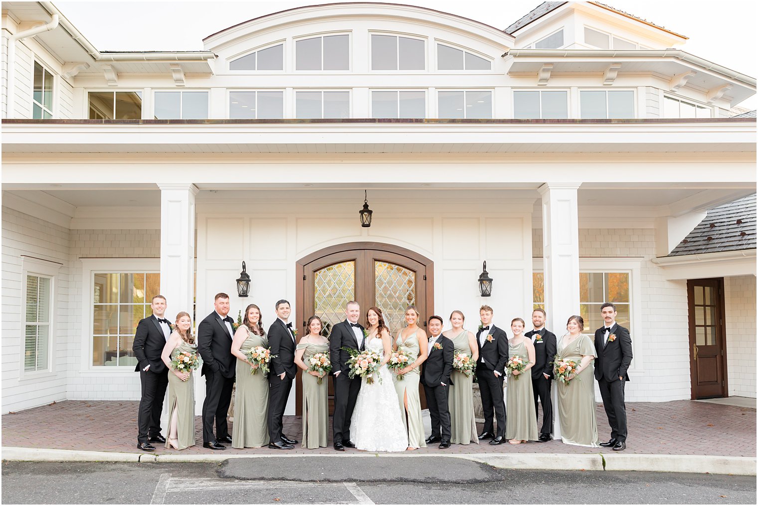 bride and groom stand with wedding party in sage green and black outside The Mill Lakeside Manor