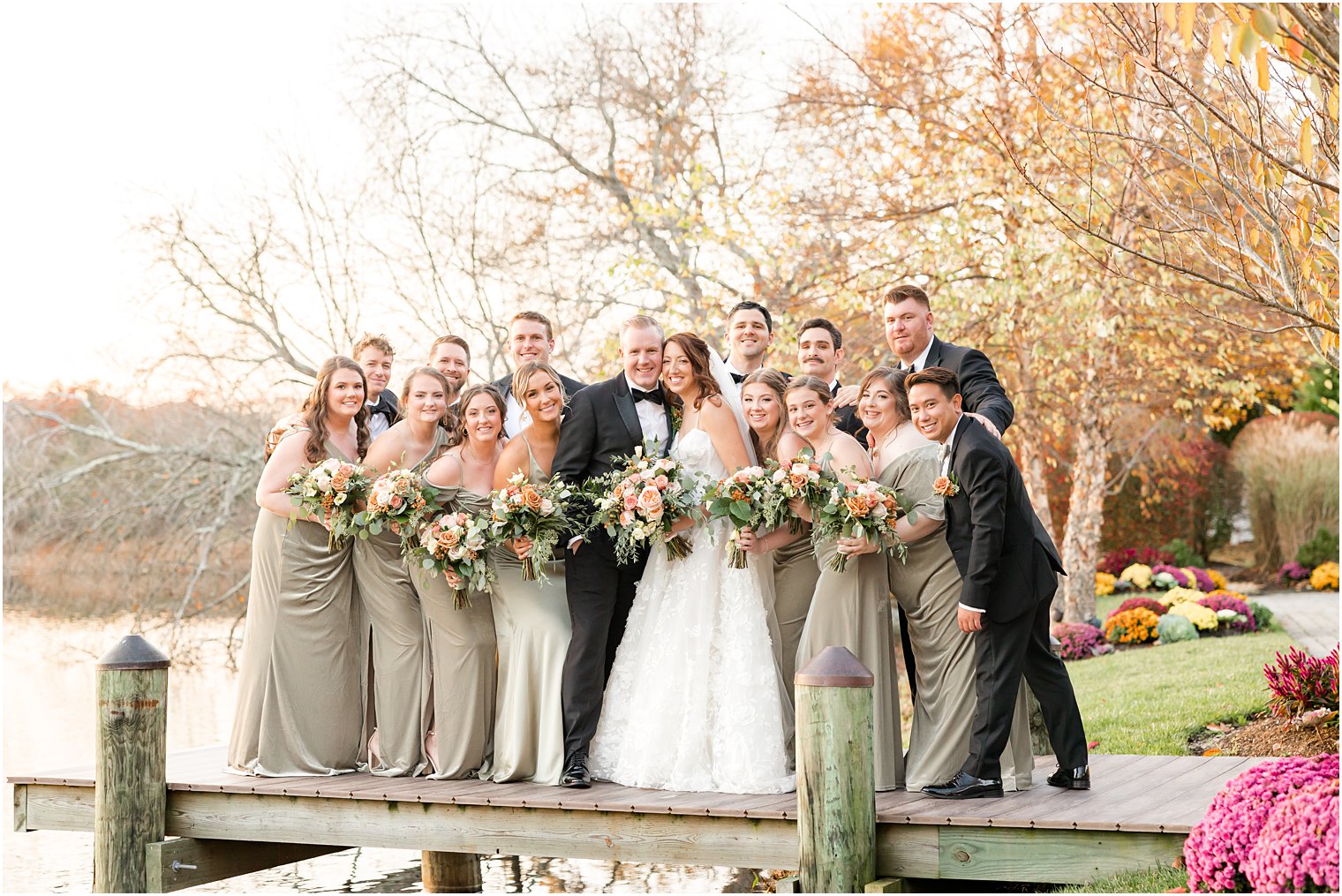 bride and groom stand on dock with bridesmaids in sage green and groomsmen in black suits 