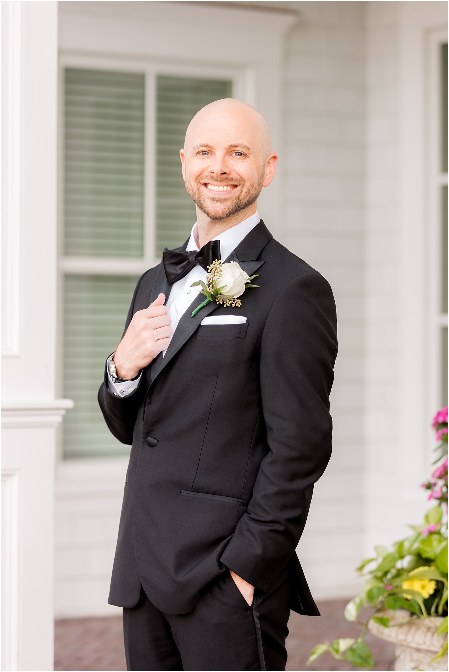 groom stands on patio at The Mill Lakeside Manor holding lapel of black tux