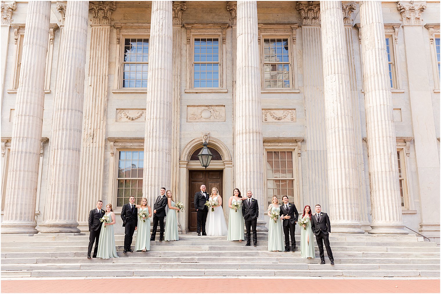 bride and groom stand on steps with wedding party in sage green and black at the First National Bank in Philly, PA