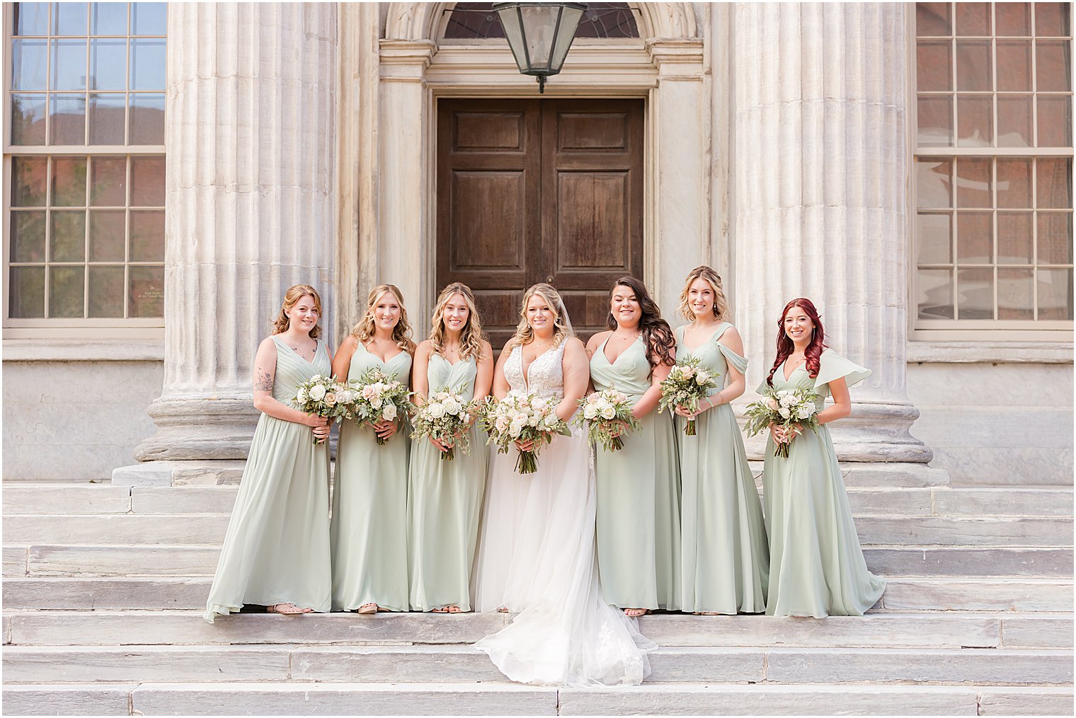 bride stands on steps of the First National Bank in Philly, PA with bridesmaids in sage green dresses 
