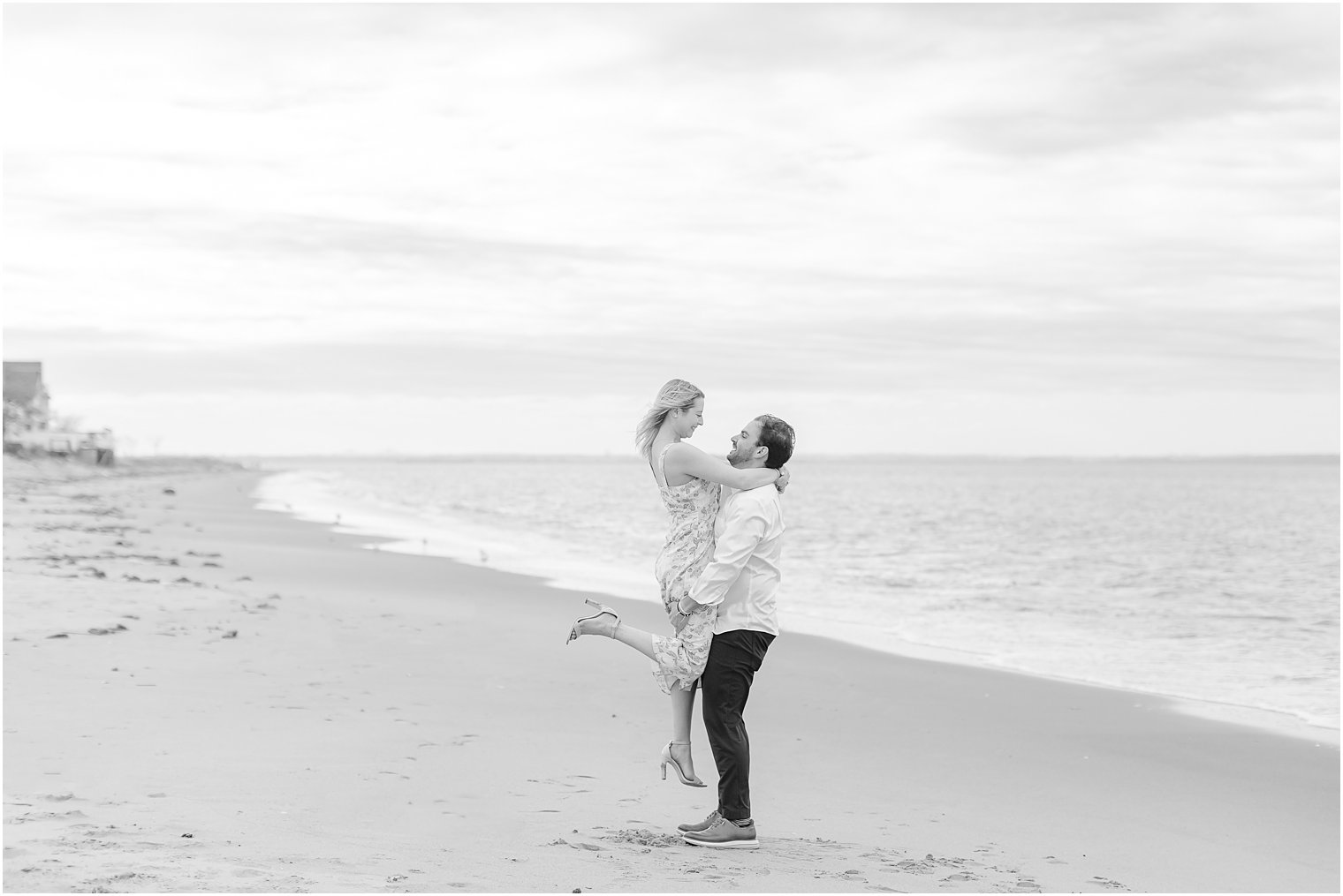 black and white portrait of couple on beach smiling while groom lifts bride up 