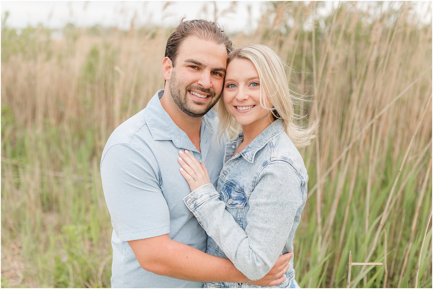 engaged couple hugs putting heads together by tall grass on beach