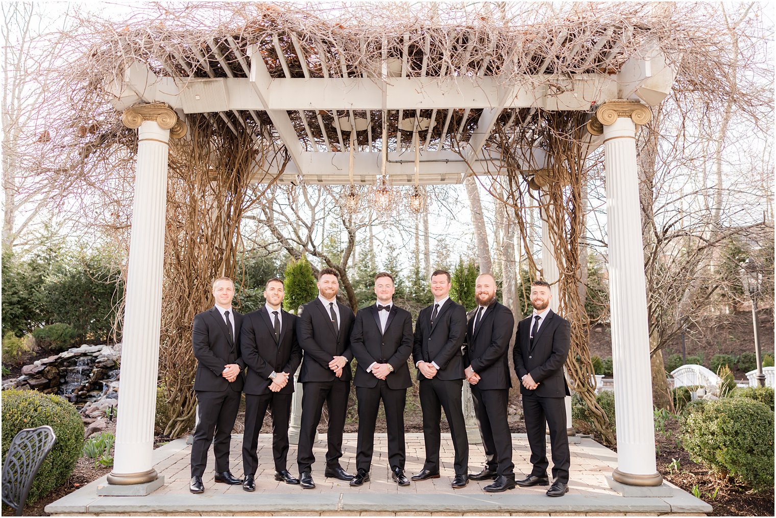 groom with groomsmen stands under arbor with pink blooms at Nanina's in the Park