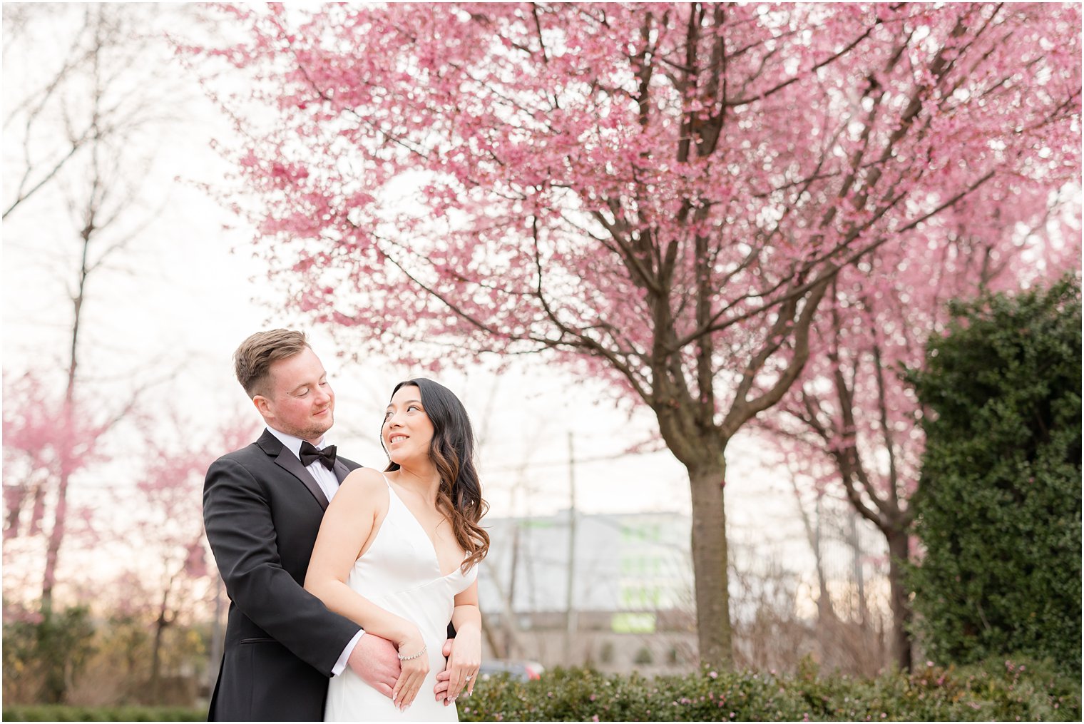 groom hugs bride from behind by pink blooms at Nanina's in the Park