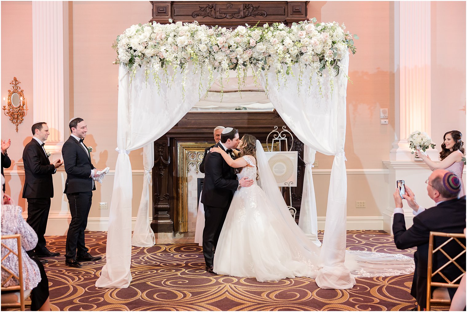newlyweds kiss under canopy with white flowers during traditional Jewish wedding at the Palace at Somerset Park