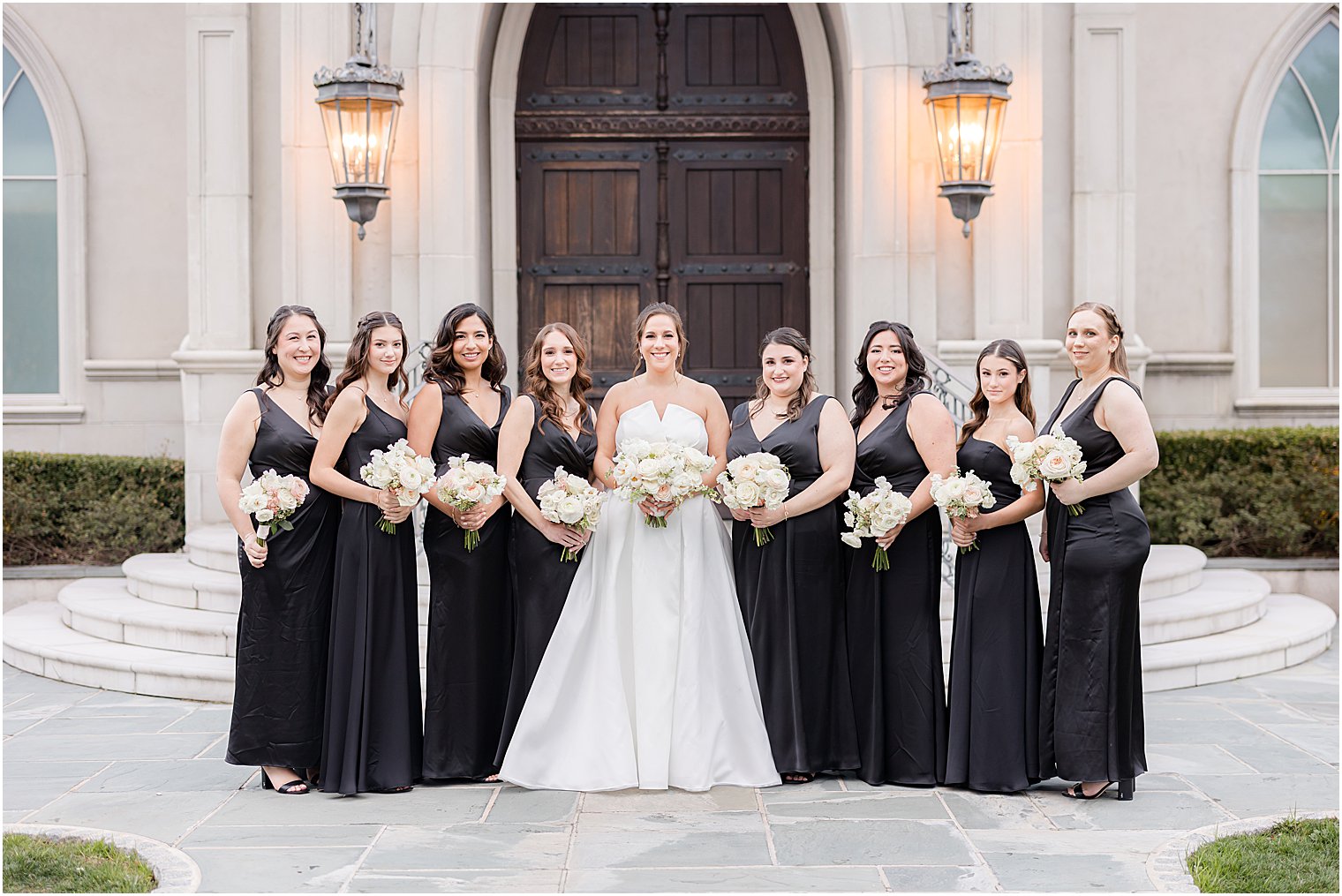 bride holds bouquet of white flowers with bridesmaids in black gowns